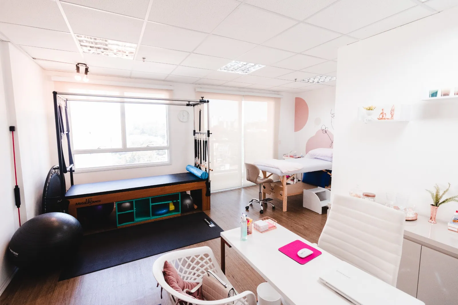 Sala ampla e iluminada com aparelho de pilates, maca e mesa de consulta